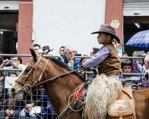 A young boy dressed in gaucho clothing to include cowboy hat, leather vest, and alpaca chaps as he rides his work horse