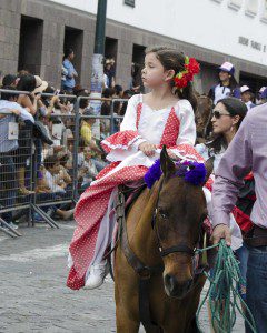 Young lady in red and pink colonial style dress rides a horse while led by another person.