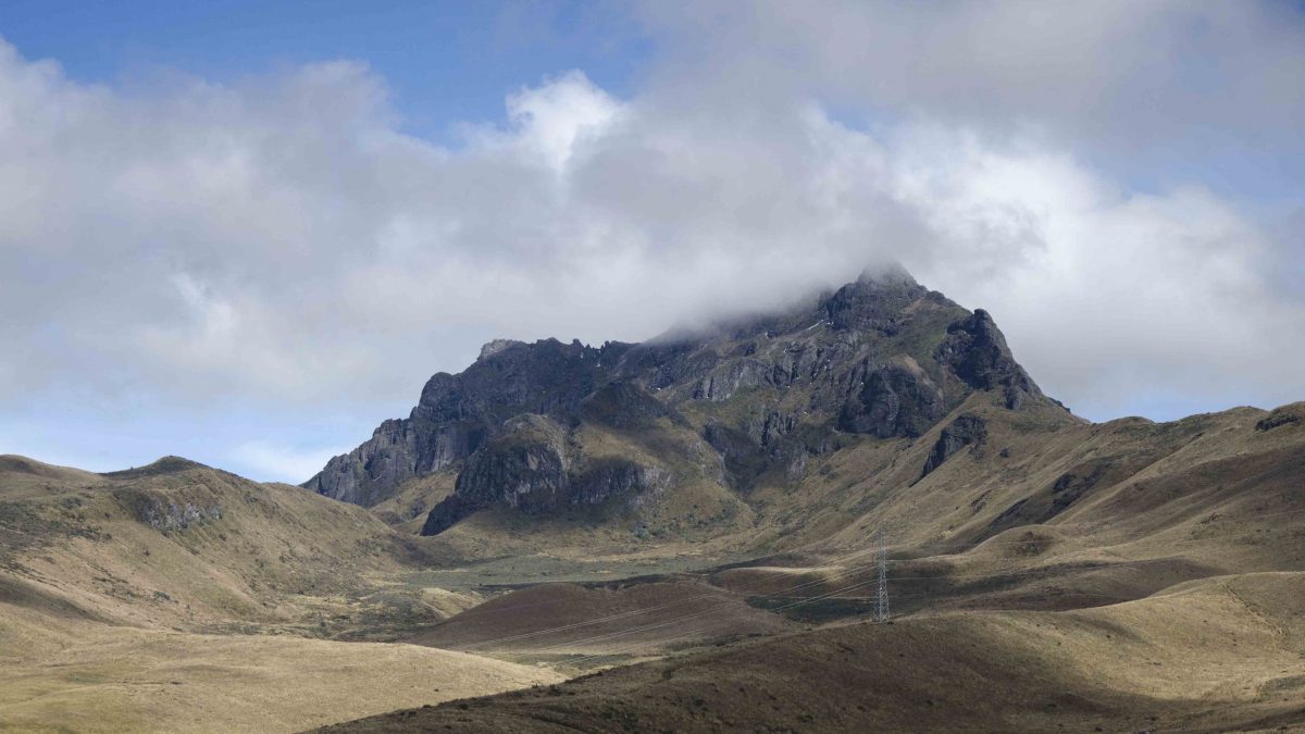 Laderas de paramo llevan la vista al pico rocoso llamado Rucu Pichincha, que se encuentra bajo un cielo azul parcialmente nublado