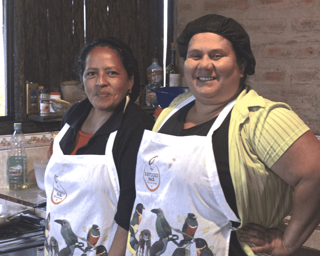 Dos mujeres sonrientes, vestidas con delantales con muchas imágenes de pájaros, están sonrientes en una cocina.