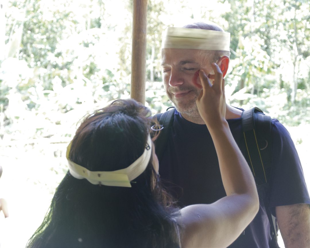 Un turista alto, blanco y masculino tiene la cara pintada con marcas rojas por una mujer Huaorani.