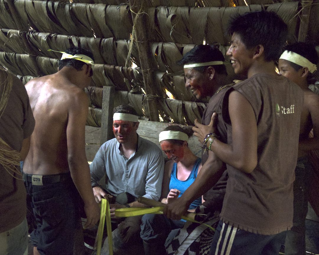 A white couple sits in the center holding hands and are surrounded by Huaorani men tying their hands together in a symbolic marriage