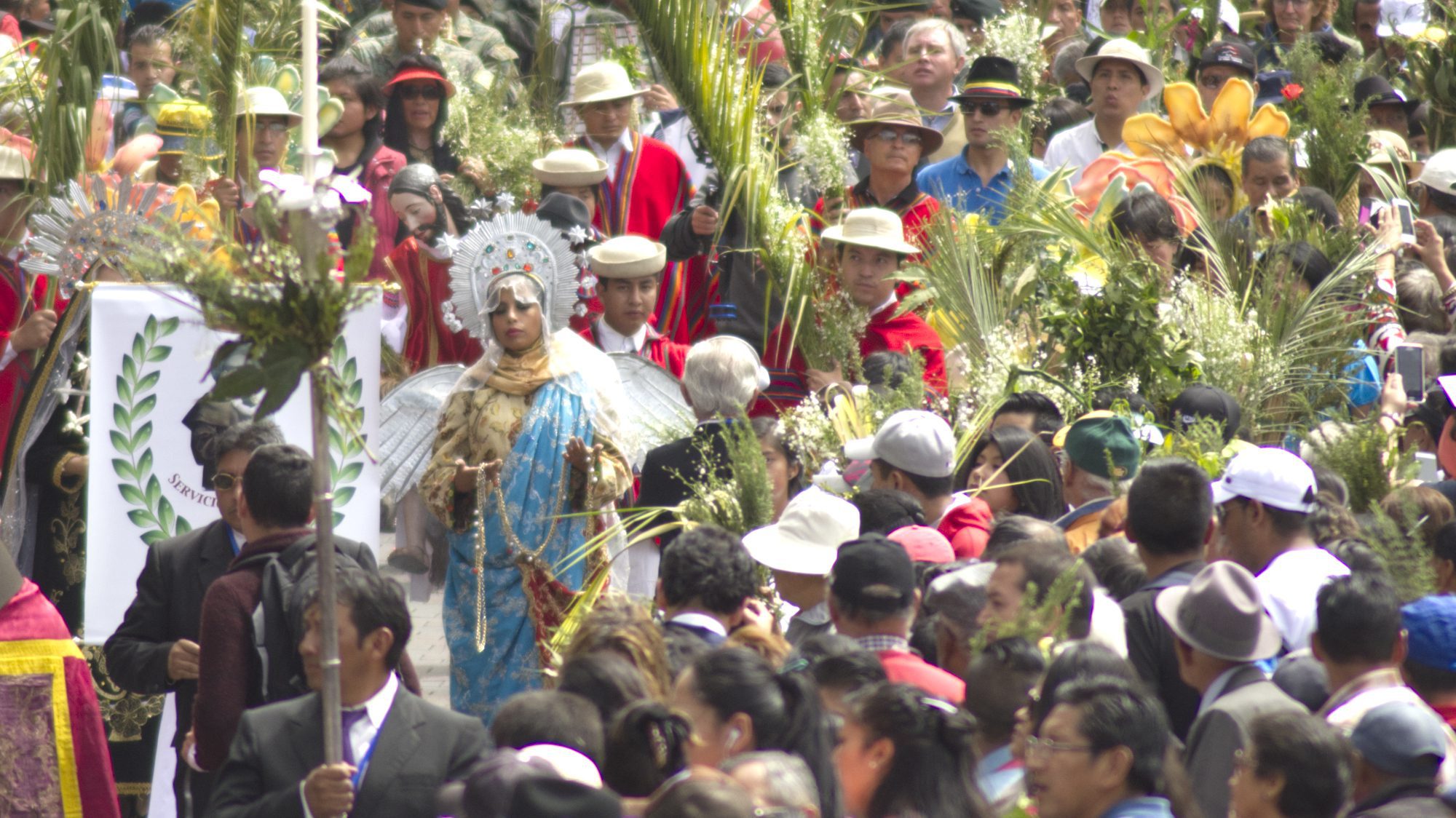 The Virgin of Quito walks along a sidewalk full of people holding bouquets for Palm Sunday