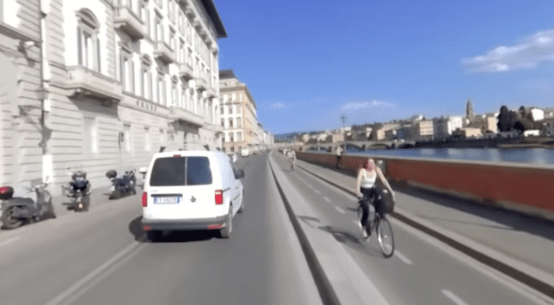 A woman on a bicycle comes toward the viewer in a bike lane to the right while we follow a white van on the main road ahead