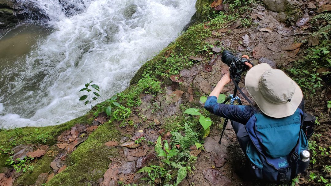 Una mujer se agacha sobre el trípode de una cámara mientras encuadra una foto de una cascada