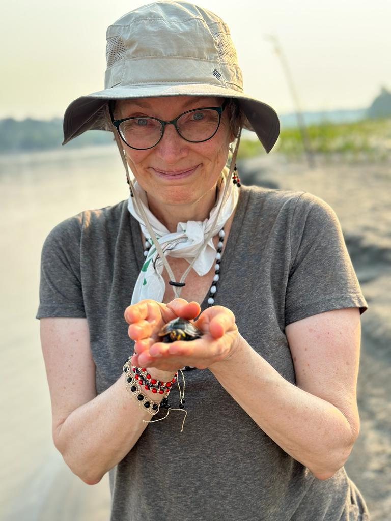 Women dressed for the outdoors with sunhat, grey t-shirt, and white bandana holds a small turtle in her cupped hands