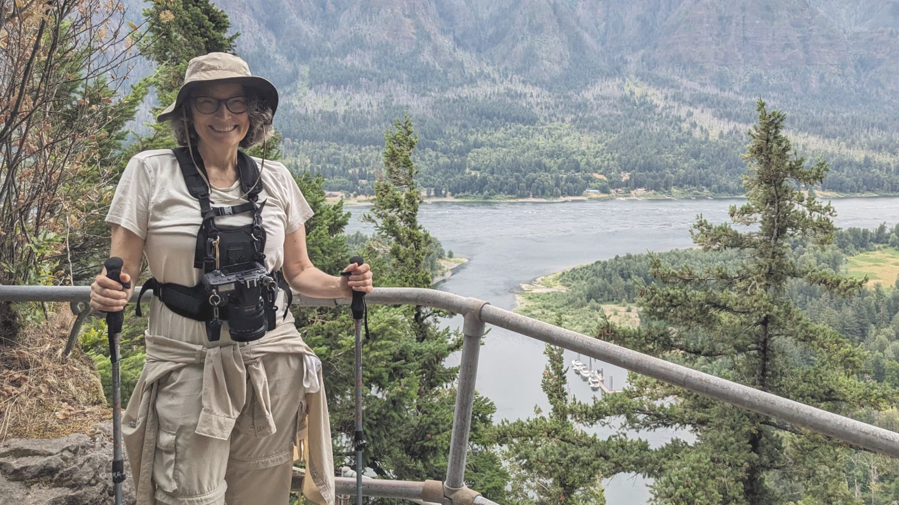 A woman dressed in hiking gear and wearing a camera harness holds hiking poles while posing in front of the Columbia River from a high trail.