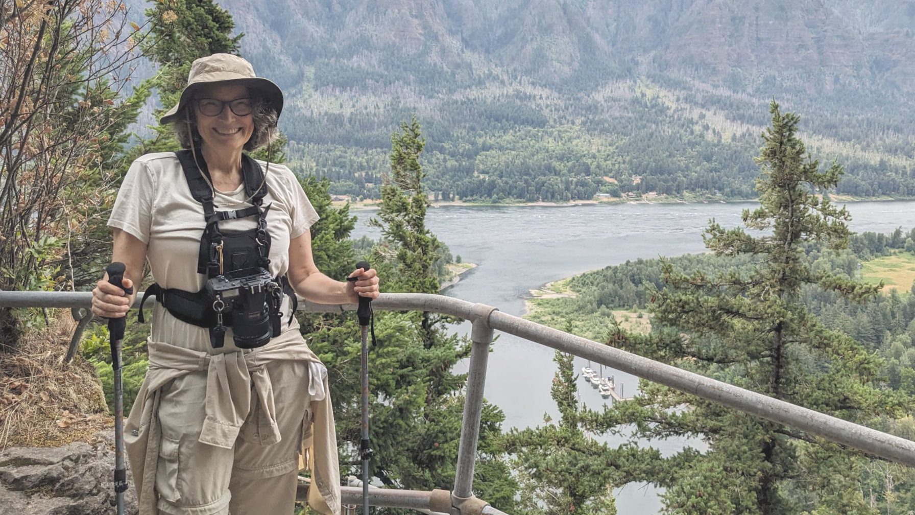 Una mujer vestida con equipo de senderismo y con un arnés de cámara sujeta unos bastones de senderismo mientras posa frente al río Columbia desde un sendero elevado.