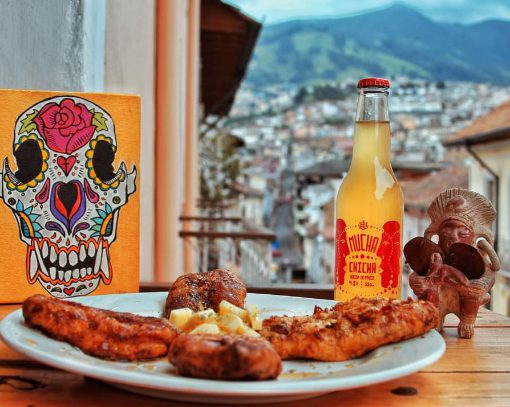 Primer plano: delicioso plato de comida, botella de chicha y arte ecuatoriano; fondo: vista de Quito.