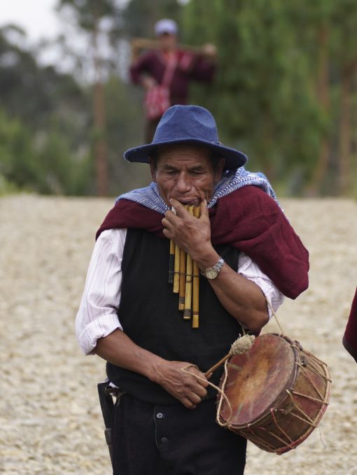 A man dressed in traditional costume plays pipes and carries a drum as he walks along a graveled road.