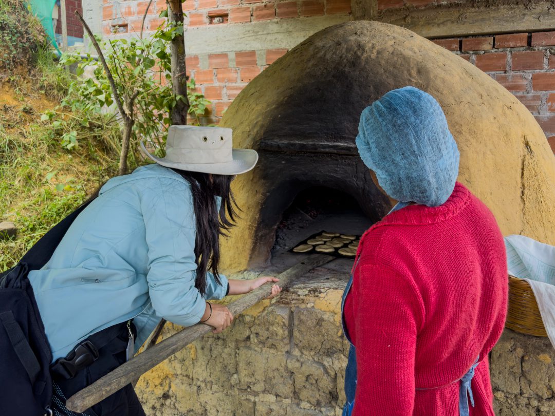 A woman places a tray of dough into a wood burning oven while another looks on.