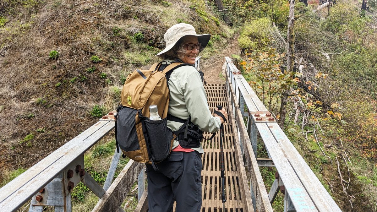 A woman in hiking gear and a a Wy'east Ultralight Backpack looks back as she walks across a bridge.