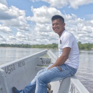 Edgar Noteno of Kichwa Lodge sits on the each of a motorized canoe on the wide Aguarico River with a sliver of green jungle visible under a stunning blue sky full of white clouds