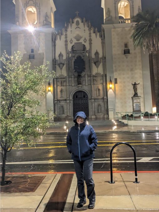 Angie standing in front of the cathedral in Tucson, Arizona wearing her Craft Sportswear Women's CORE Explore Shell Jacket while it pours with rain.<br />
