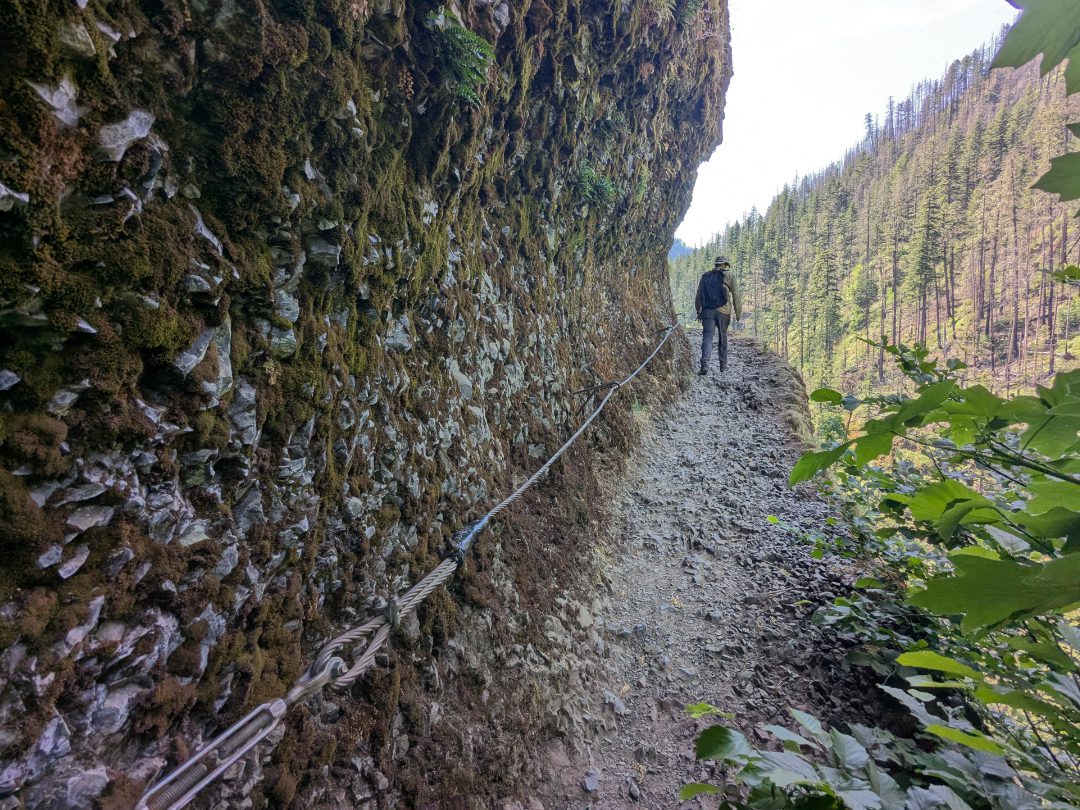 Trail to Punch Bowl Falls A lone hiker climbs a narrow trail on the edge of a cliff, cables along the mountainside to the left, a drop off and valley view to the right