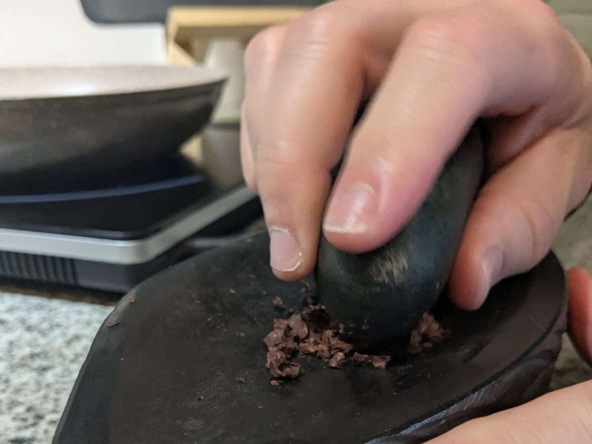 Close-up of a hand holding a mortar while grinding cacao beans on a flat pestle.