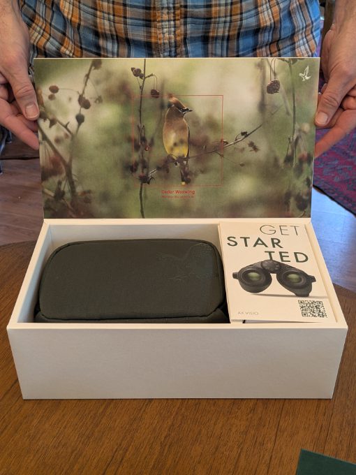 A pair of hands holds the top of a box open to display its contents: a set of AX Visio Smart Binoculars