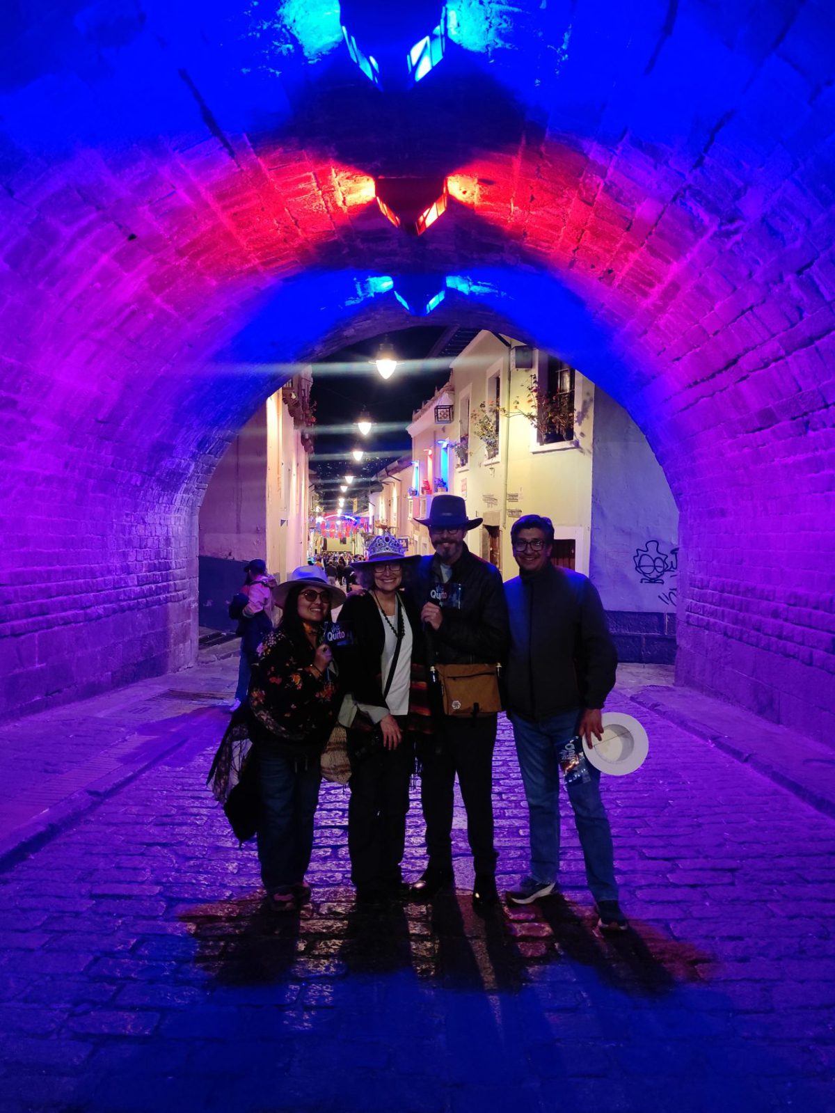 Four friends stand beneath a tunnel lit in reds, blues, and purples in celebration of the Fiestas de Quito in La Ronda.