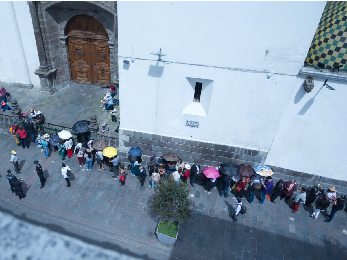 People lined up against the cathedral wall, holding umbrellas to protect themselves from the equatorial sun, wait to enter the Arrastre de Caudas