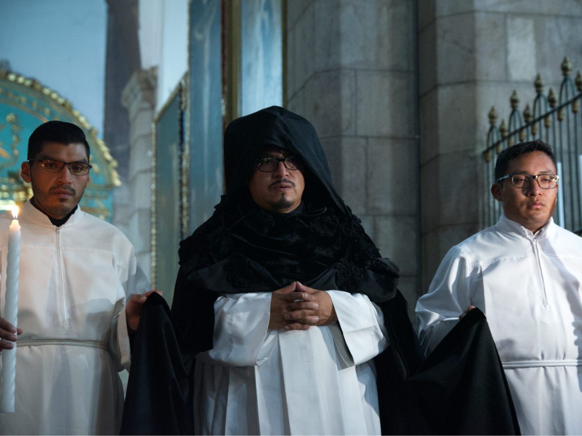 Second priest, clasping his hands, in hooded black robes walking with two acolytes holding candles, Arrastre de Caudas Quito