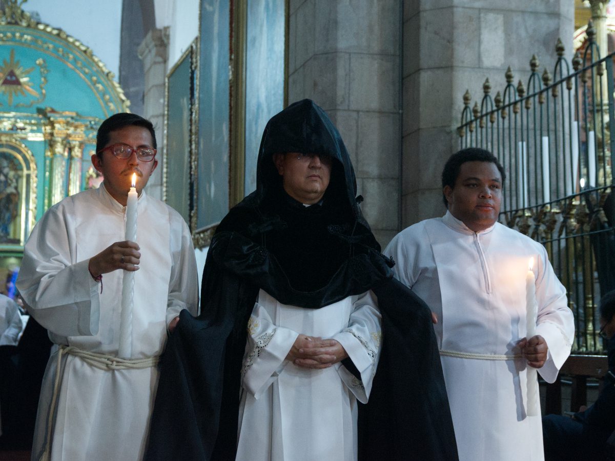 Canon in black cowl guided by two acolytes with candles during Arrastre de Caudas in Quito