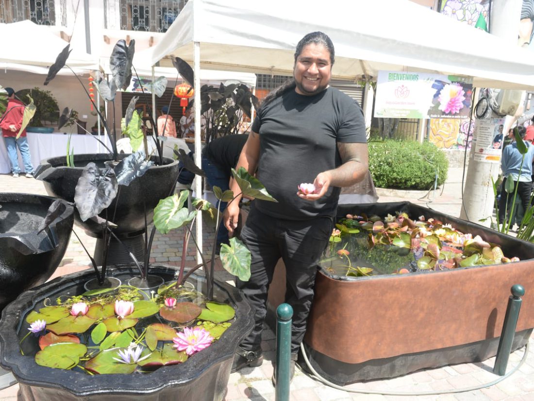Un hombre sonriente sostiene una pequeña flor rosada mientras posa junto a estanques ornamentales de jardín con nenúfares en flor.