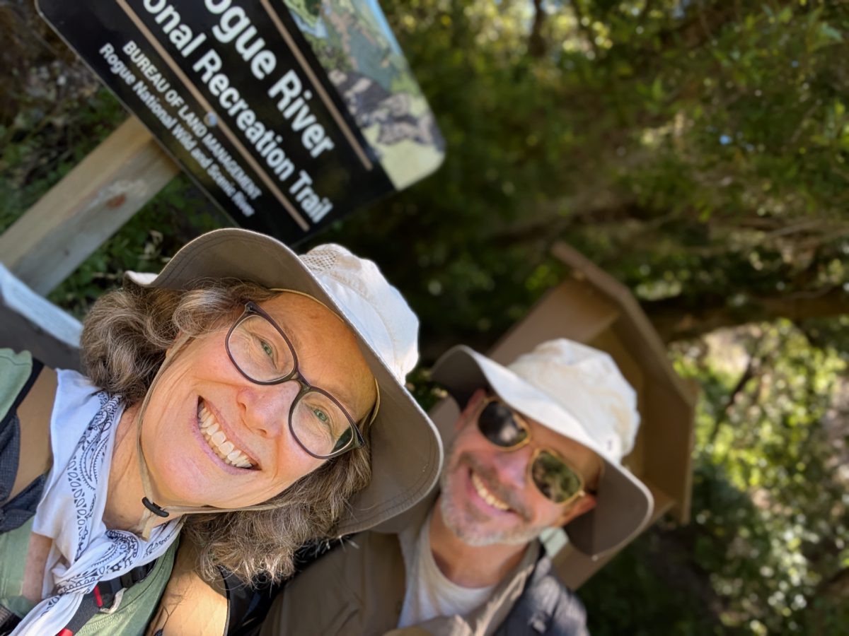 Two smiling hikers take a selfie in front of the Lower Rogue River Trail Sign.