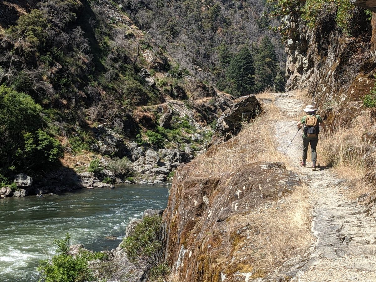 A hiker on a narrow trail on a cliff edge that overlooks a dark blue green river.