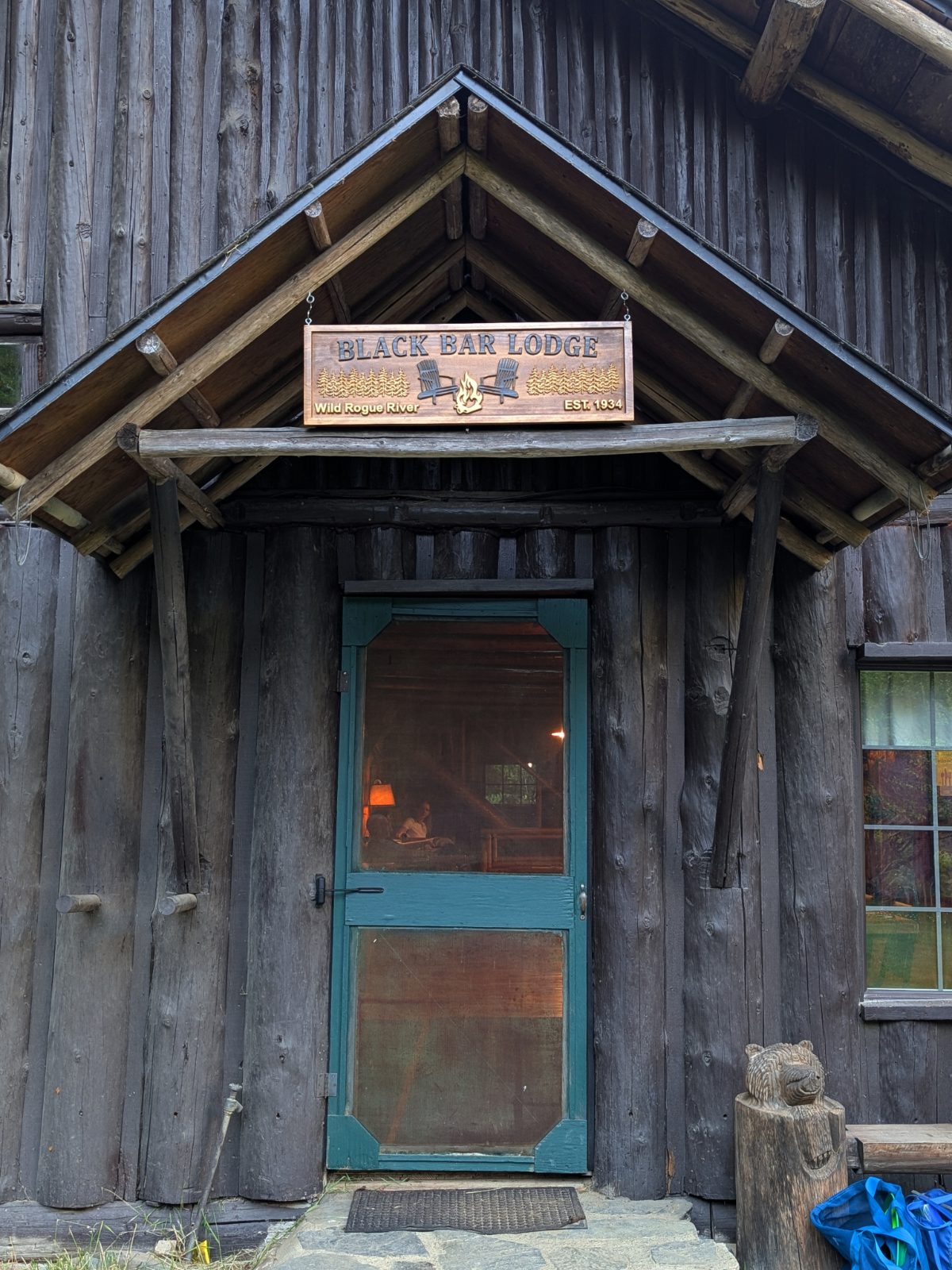 A teal blue screen door under a rustic carved sign saying Black Bar Lodge, Wild Rogue River, Est. 1934.
