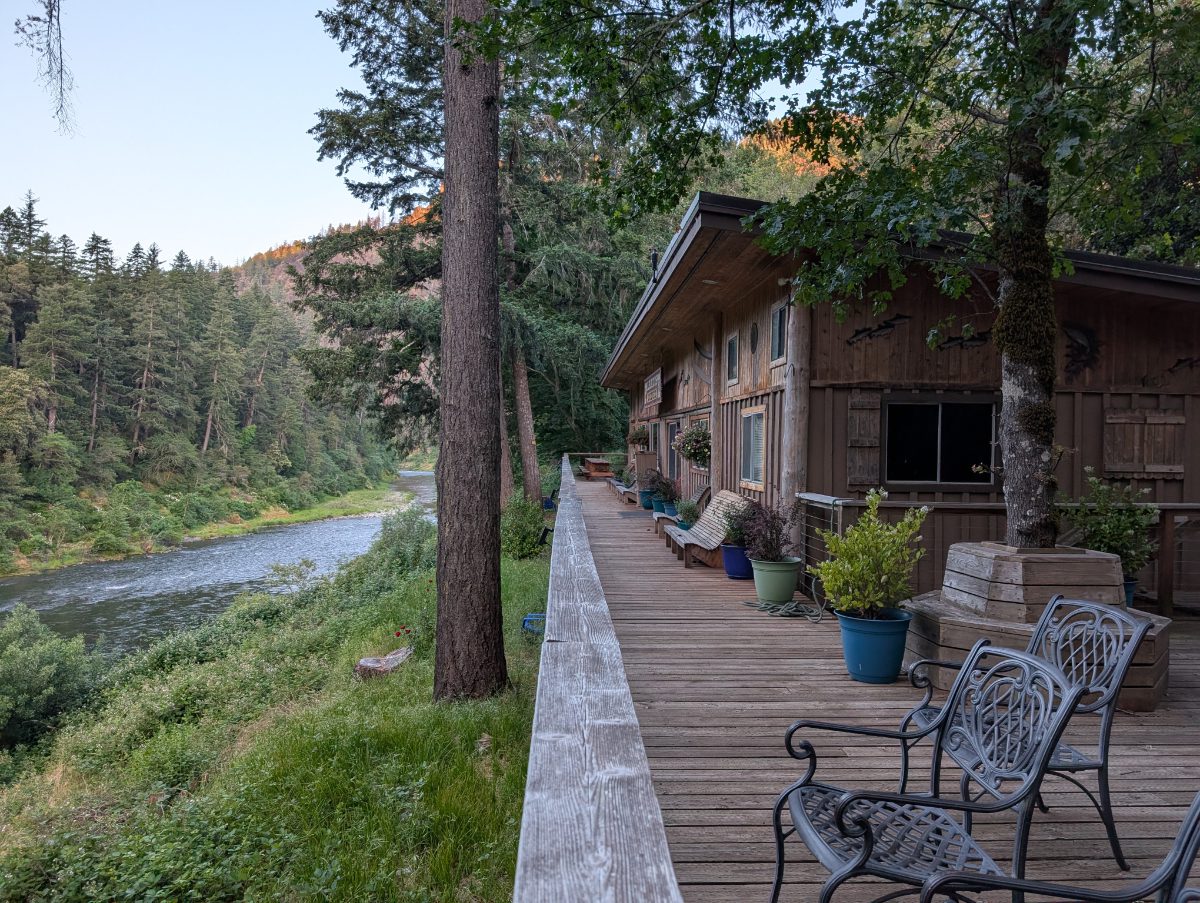 A long outdoor patio surrounds the wooden building of the dining room, kitchen, and hang out space at Paradise Lodge, all with views of the Rogue River.