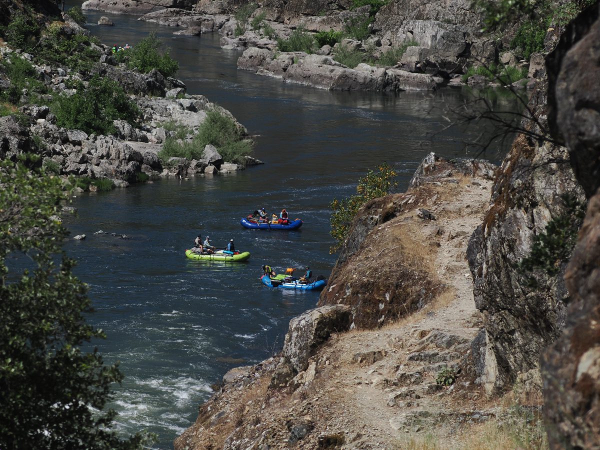 Raft or Hike the Rogue River? This image makes the choice apparent with a shot of rafts going down the lower Rogue and a narrow rocky path in full sun hugging the cliff's edge.