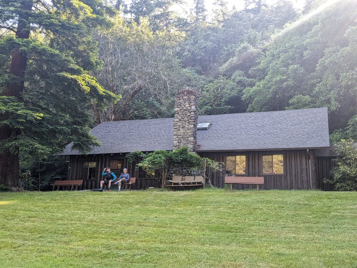 Two people sit on a bench outside a log cabin with stone chimney