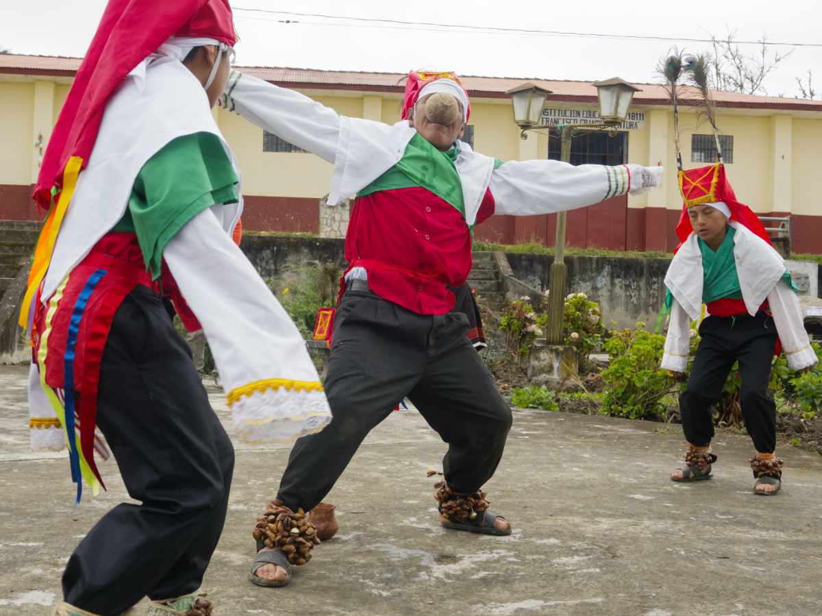 Three Chuquiaj dancers in Cuemal, Peru: an elder with arms outstretched and a gourd cup in his mouth, flanked by two young boys dancing around him.