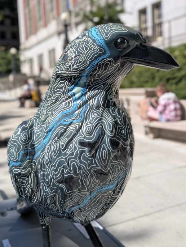 From the front, a close-up of As the Crow Flies shows a turquoise blue river flowing down its right side. 