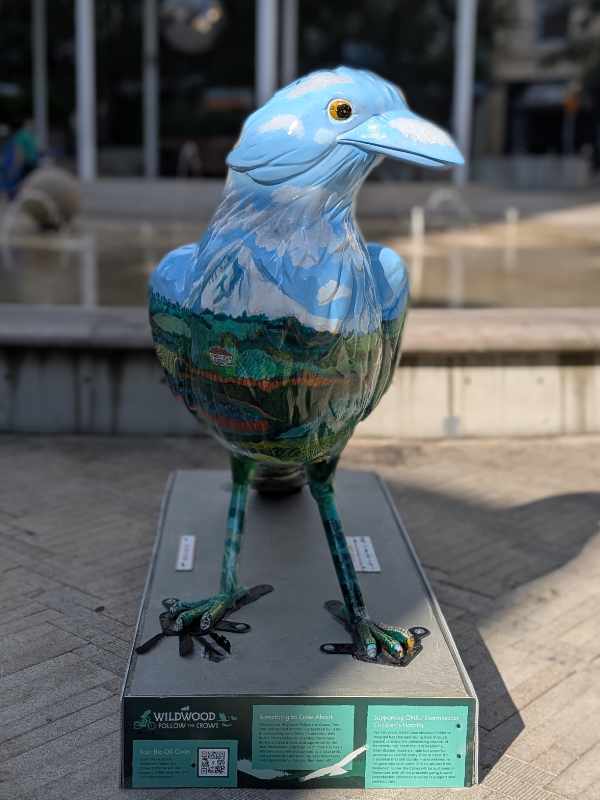 Front of the crow sculpture Flying Fields Forever, note the upper half is painted sky blue shot with white clouds behind a snow covered Mount Hood, the lower half with green trees and farm fields.