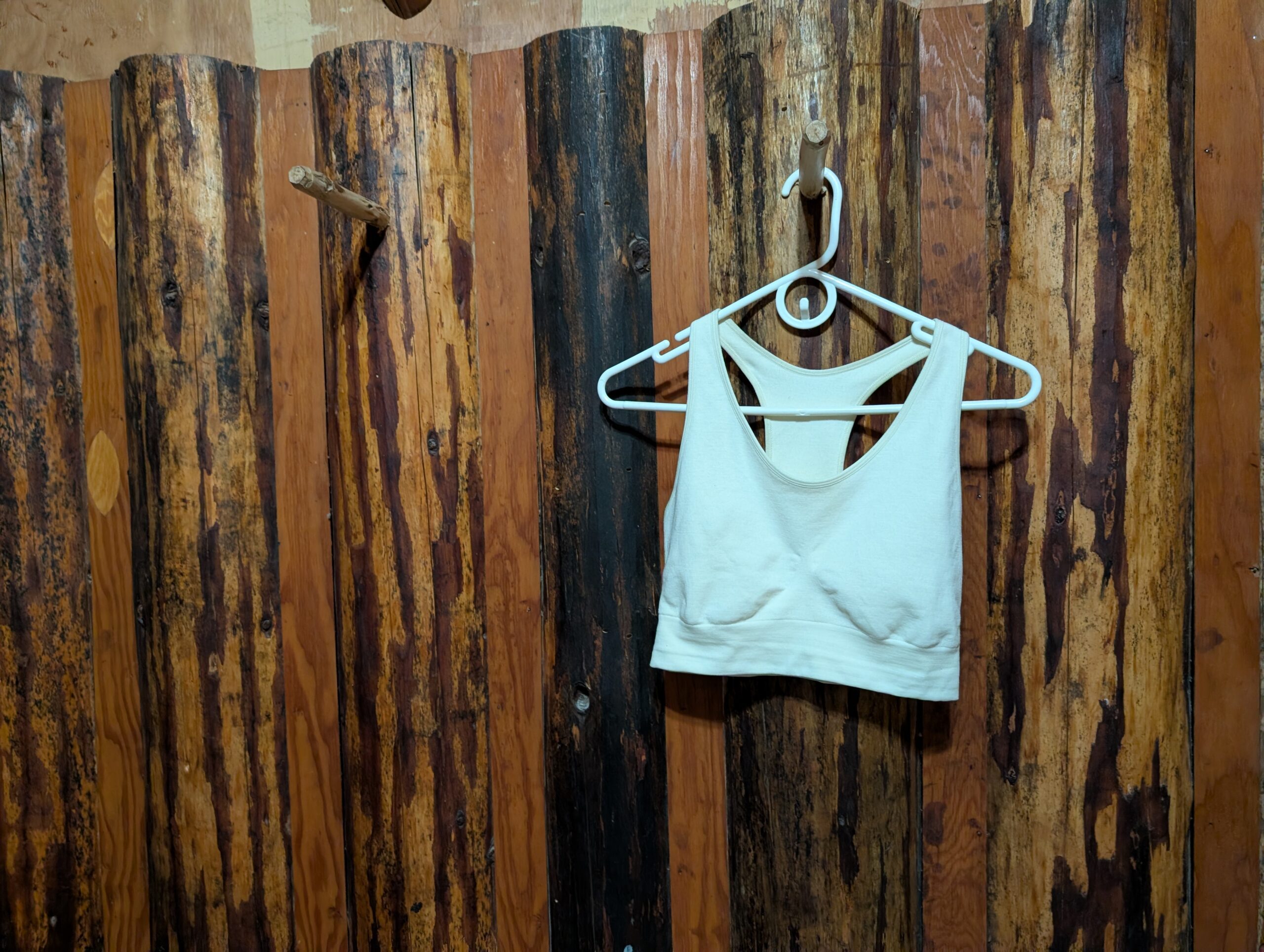 A cream colored merino wool bra on a white plastic hanger hangs from a peg on a rustic log wall.