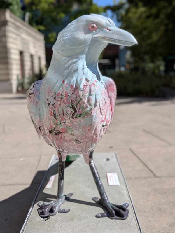 A front view of the crow statue, Sakura Hanami reveals pale pink cherry blossoms against a pale blue sky.