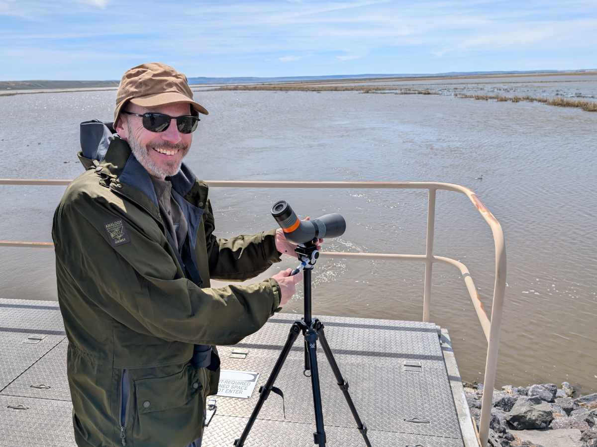Scott smiles happily at the camera while getting ready to look through a Maven spotting scope at water fowl in the extreme distance.